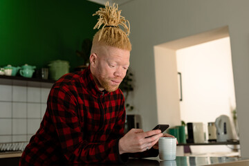 Thoughtful albino african american man with dreadlocks using smartphone and drinking coffee