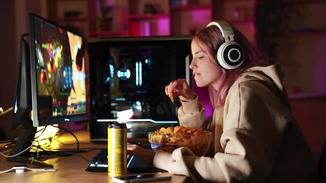 Side view of smiling gamer girl watching a video game match on a computer and eating chips