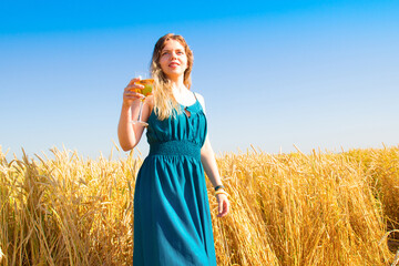 Obraz premium Lovely blonde woman in a turquoise dress with a glass of champagne in her hand against the background of a golden wheat field and blue sky on a autumn sunny day