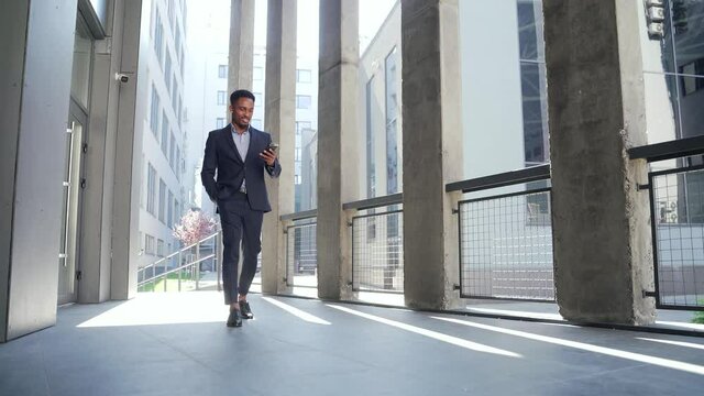 successful young confident African American businessman moving with cellphone in hands against modern office building background downtown black business man walks using a mobile phone in formal suit