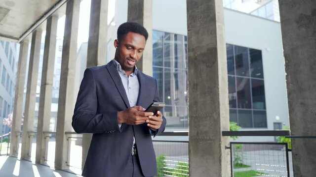 African American employee on urban background office building texting Messages, read good news mobile phone. Outside. Business man standing a formal suit messaging using Smartphone cellphone. outdoors