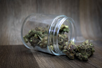 Purple marijuana buds spilling out of a glass jar onto a dark grey wooden background.