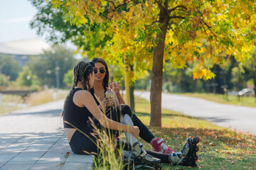 Two girls sit in a park while taking a break from rollerskating drinking smoothie and gossiping. Girls bonding concept.