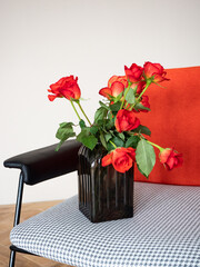 Red roses in a dark glass vase next to a retro red-black chair against a light wall and wooden floor in natural morning light