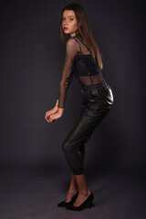 Full-length fashion portrait of an attractive female wearing leather pants and see-through shirt while posing on dark background