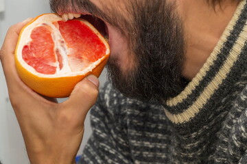 A bearded man in a sweater bites into a grapefruit