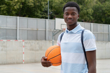 African American student back to school. Portrait of a Black teenager boy outside high school with a basketball ball in the football court.