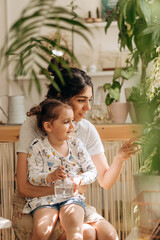 Happy mixed race family woman with her little daughter is spraying houseplants with water from a...