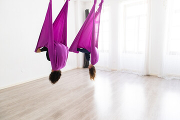 two young women friends practicing aerial yoga fly yoga in studio with hammock having fun moving...