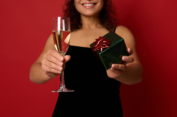 Soft focus on champagne flute with sparkling wine and box wrapped in glitter gift paper in the hands of pretty woman with beautiful toothy smile. Christmas concepts on red background with copy space