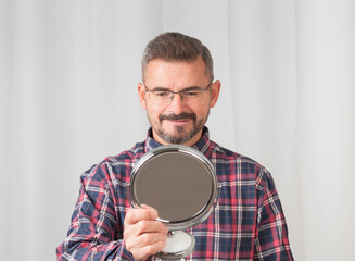 Senior man looking on portable mirror on gray background