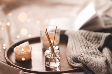 Home perfume in glass bottle with wood sticks, scented burn candles, open paper book and knit wool...