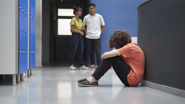 Sad teenager sitting alone on floor victim of bullying while classmates ignore him. Bullied student crying and suffering depression. Problem in Education
