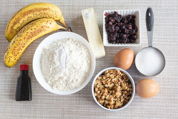 Banana bread ingredients. On the table are bananas, eggs, flour, butter, walnuts, dried cranberries, vanilla essence. Top view, daylight, neutral background.  Homemade baking. 