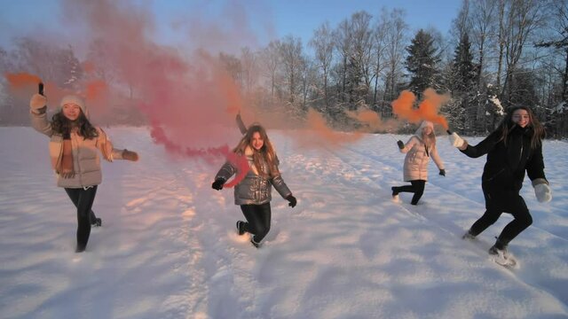 Girls friends run across the winter field with colored smoke.