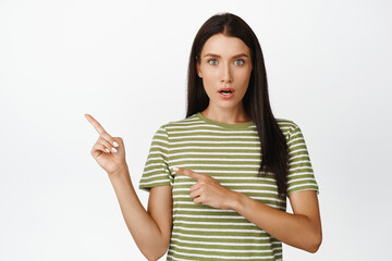 Amazed woman pointing left, looking surprised, showing smth interesting, standing over white...
