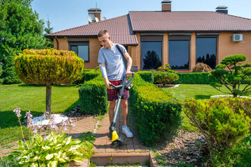 Young man with amputated arm and prosthesis removing weeds on the sidewalk of a backyard garden...