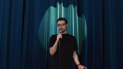 A European man stand-up comedian on stage with a microphone in his hands says goodbye to the audience, leaves against the background of blue curtains.