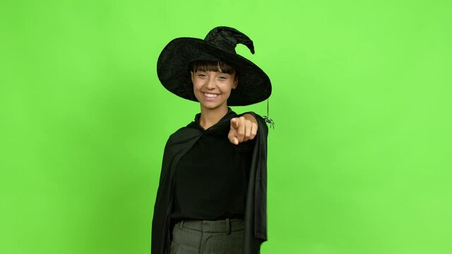 Young woman wearing witch hat points finger at you with a confident expression over isolated background. Green screen chroma key