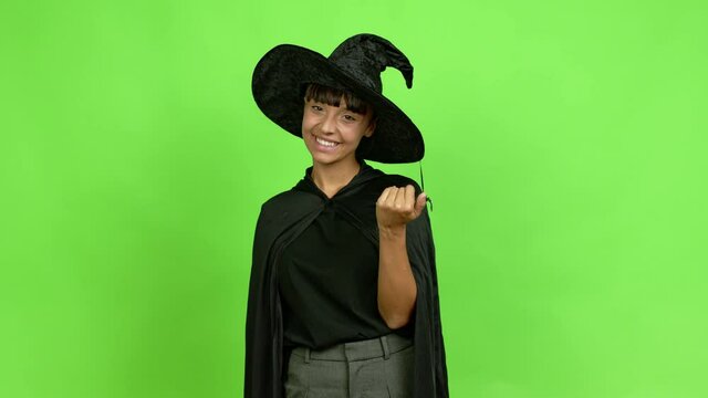 Young woman wearing witch hat presenting and inviting to come with hand over isolated background. Green screen chroma key
