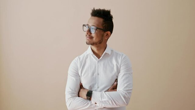 Young man wearing glasses and white shirt turning head side by side thinking and generating ideas in his mind crossed arms on beige background