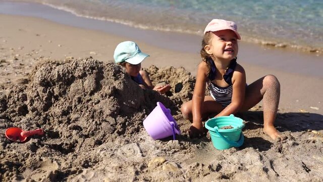 Children girls playing with sand at the beach in summer day. Kids building sand sculptures with shovels and buckets on coast sea. Fun family vacation concept