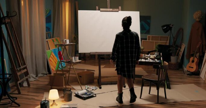 Dark art studio shadow of woman approaches large easel with white canvas around mess of paints, brushes, girl touches structure, checks unevenness of backing from painting she looks at