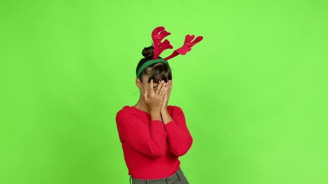 Young woman wearing christmas hat covering eyes by hands and looking through the fingers over isolated background. Green screen chroma key
