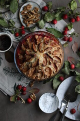 Apple charlotte sprinkled with powdered sugar in a glass baking dish. Autumn pastries on the table with a tablecloth and branches with leaves.