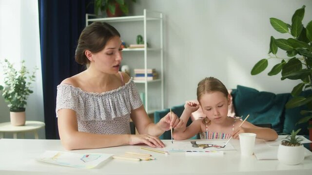 Young mother teaching her daughter to draw. Lovely babysitter drawing with little girl at home, using watercolor. Motherhood and happy childhood concept. Preschool children education.