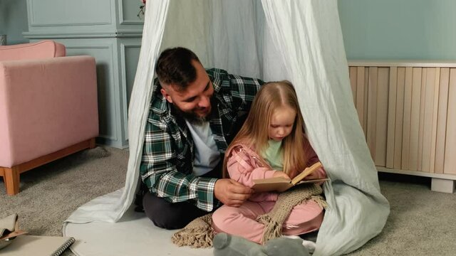 Happy family adult parent dad and small daughter having fun reading book lying on floor at home. Cute child girl learning how to read