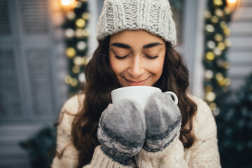 Young beautiful woman wearing knitted hat and white sweater sitting on porch of holiday decorated house with festive lights. Christmas decorations.
