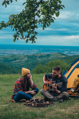 Couple of campers sitting near the camp fire and a tent and playing guitar