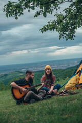 Couple of campers sitting near the camp fire and a tent and playing guitar