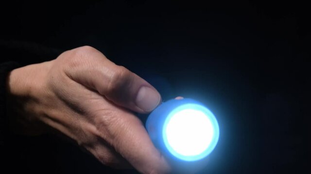 Hand with a led flashlight shining in the darkness with blue beam into the camera, spy and investigation concept