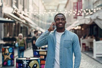 Fototapeta premium Attractive joyful afro-american guy with stylish beard has phone conversation during walking on the street at summer.