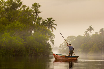 Misty morning and a man, fisherman on the boat on the river with a wooden stick moving between palm...