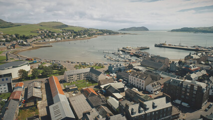 Harbor town cityscape aerial. Seascape with yachts, ships at urban street with road. Cars drive at traffic highway. Modern buildings at ocean shore of Campbeltown, Scotland, Europe rise-up drone shot