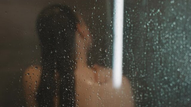 Woman washing herself in the shower behind the glass.