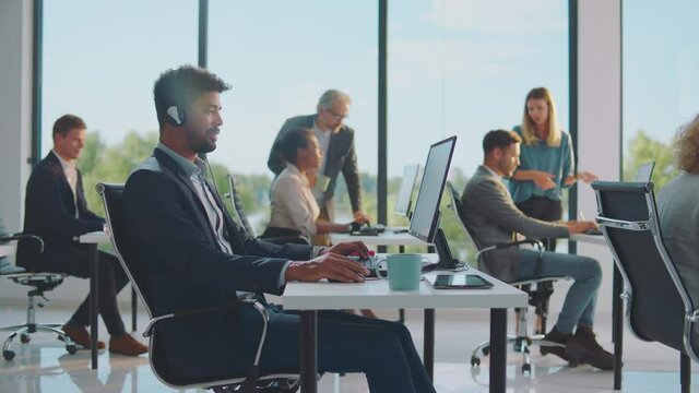 Working in noisy office, using headset for phone call to client. Colleagues gesturing while discussing. Modern open space office interior.