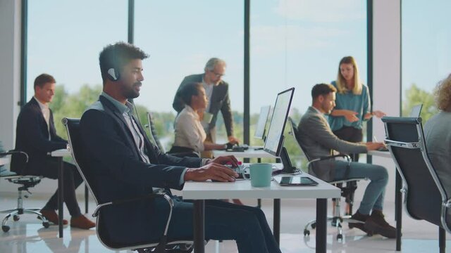 Working in modestly furnished room with large windows. Mixed race businessman using headset for communication. Modern open space office interior.
