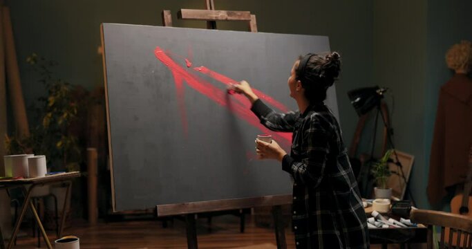 An entertaining artistic soul, woman stands in front of canvas painted gray, holding can of red paint and brush, pursuing abstract vision, splashing on painting, dragging color in different directions