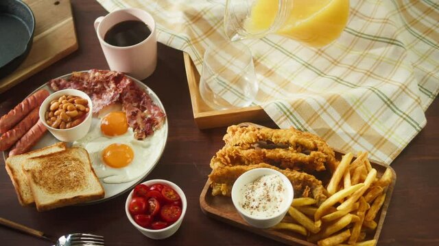 Traditional full English breakfast close-up. National British cuisine, fried eggs with bacon and sausages, toasts, fish and chips, baked beans, top view. Pouring orange juice. American pub or cafe. 