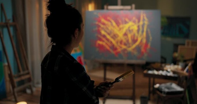 A shadow of a woman standing close to the camera, facing the canvas with masterpiece, a girl is looking at work holding a brush dirty from paint, an easel in the middle of a painting studio