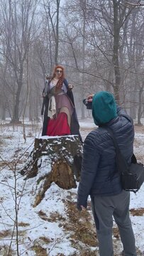 Funny woman with dreadlocks in celtic costume and with axe dancing on large stump in front of photographer. Man in hooded jacket shoots cosplayer girl with creepy makeup in snowy forest.