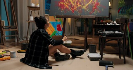 A woman is working in her studio, school, and art class on a new piece of painting related to abstractionism, modernity. The girl crouches on the floor picks up a tray and thinks about the color