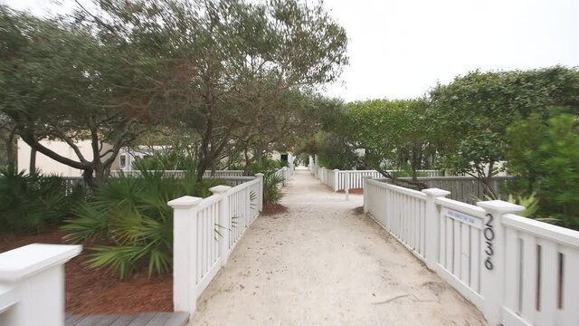 Pov point of view walking in Seaside, Florida by white beach wooden architecture house home real estate rental timeshare property with path alley by green landscaping shrubs, bushes sidewalk