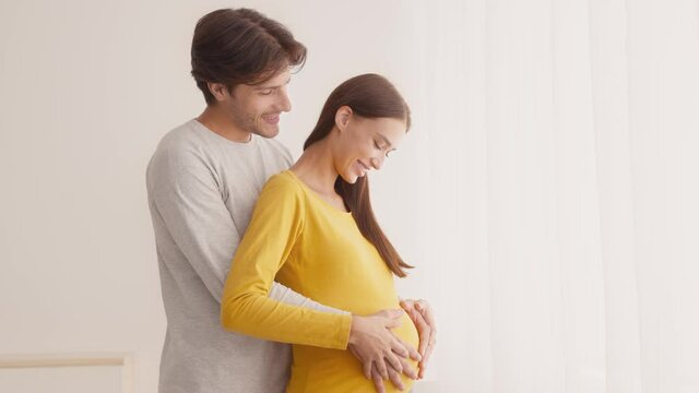 Loving man hugging his pregnant wife from behind, caressing belly,, standing near window at home, empty space