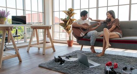 Young attractive Asian couple sitting together on a couch. Man playing guitar and woman listening and smiling in living room with glass windows background. Concept for love and couple relationship
