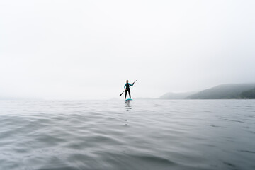 Fitness woman standing on surfboard enjoying supsurfing with paddle at endless sea water fog